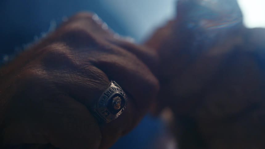 An extreme close-up of a man's hands clasped together, with a class ring catching the moody, low light