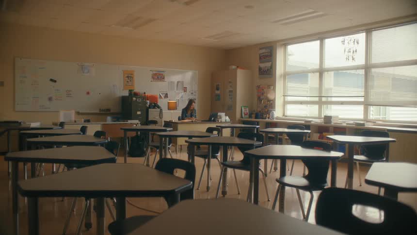 A teacher sits alone working at her desk in a large, empty classroom