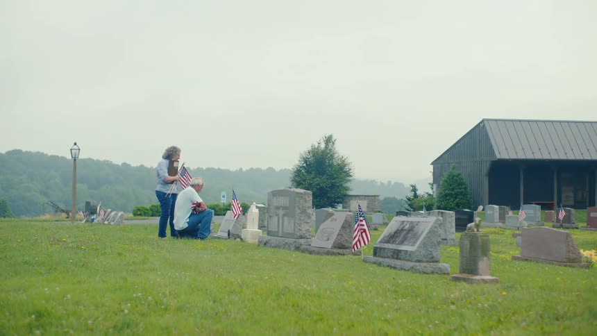 A man kneels in sorrow at a gravesite while a woman comforts him, both paying respects in a cemetery adorned with American flags
