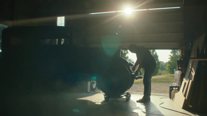 A silhouette of a man working on a classic hot rod in a dusty, sunlit garage