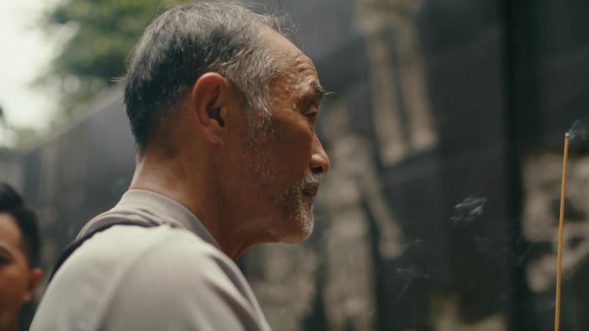 A reverent elderly Asian man contemplates peacefully while holding a smoking incense stick at a sacred temple