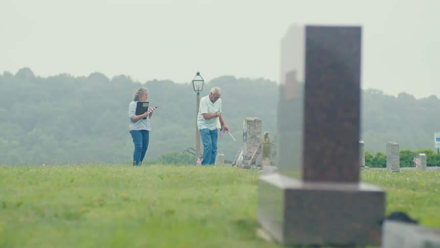 An elderly couple honors fallen veterans by walking through a cemetery and placing American flags at their headstones on an overcast day
