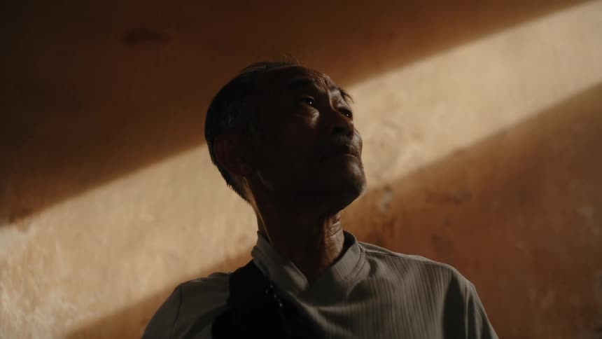 A senior Asian man with a thoughtful expression looks up towards a beam of warm sunlight in a dimly lit room