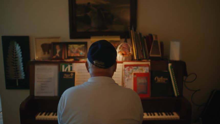 A senior man wearing a cap sits at an upright piano cluttered with sheet music and photos