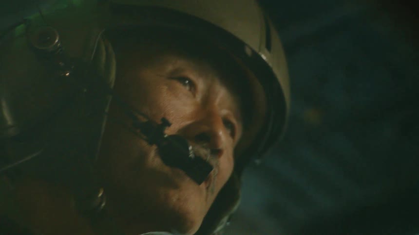 An intense close-up of an older military pilot in a helmet communicating through his headset inside a dark cockpit