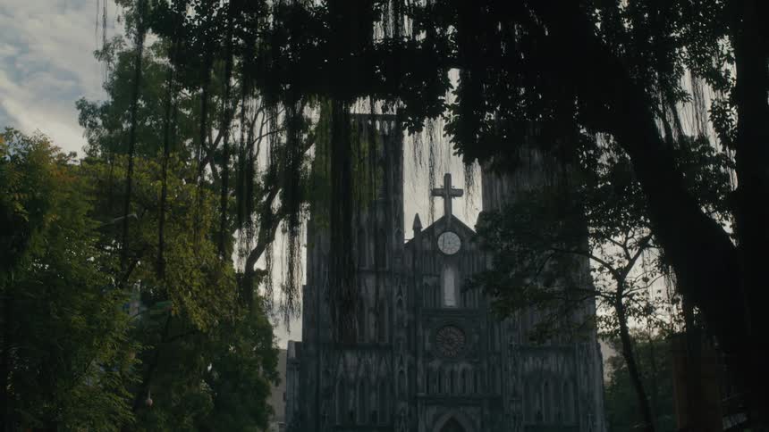 An imposing grey stone cathedral with twin bell towers is framed by the dark silhouettes of lush green trees