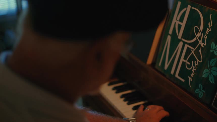 Over-the-shoulder shot of an elderly man playing a beautiful melody on an upright piano in a warm, cozy room