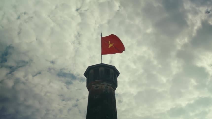 A Vietnamese flag waves in the wind atop a stone tower against a dramatic cloudy sky