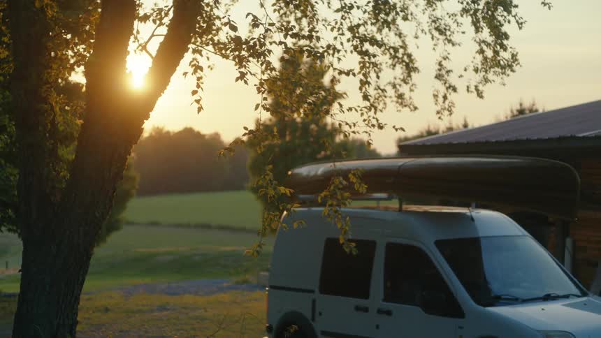 An adventure van with a canoe on its roof is parked in a peaceful rural setting at sunset