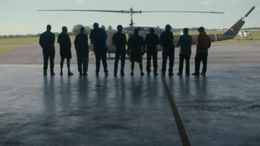 A group of men stand silhouetted in a hangar, looking out at a military helicopter on the tarmac