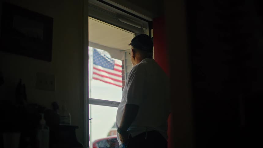 A patriotic older man stands silhouetted in a dark doorway, thoughtfully looking out at the American flag waving in the wind