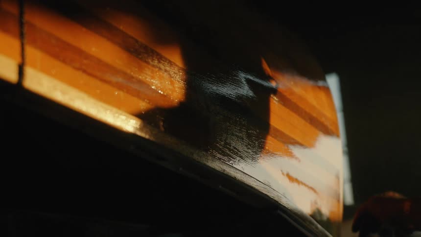 A craftsman's silhouetted hand gently applies varnish to the side of a beautiful wooden canoe in a sunlit workshop