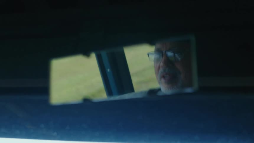 A close-up shot of a senior man's reflection in a rearview mirror as he drives through the countryside