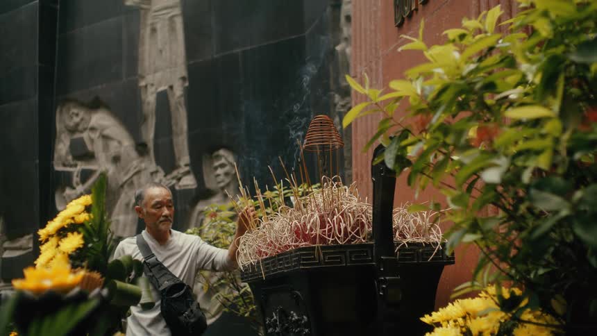 An elderly Asian man places a smoking incense stick in a censer at a serene temple, surrounded by offerings of bright yellow flowers