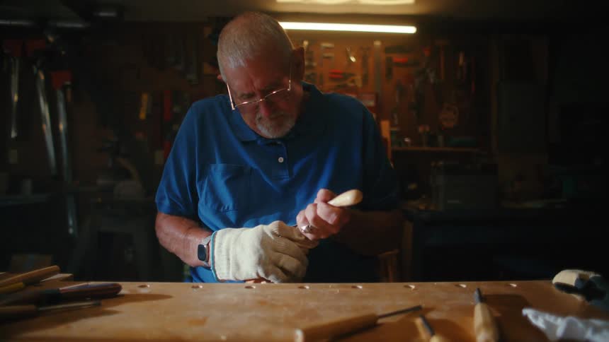 A senior craftsman with glasses intently carves a piece of wood with a whittling knife at his workbench in a dimly lit workshop