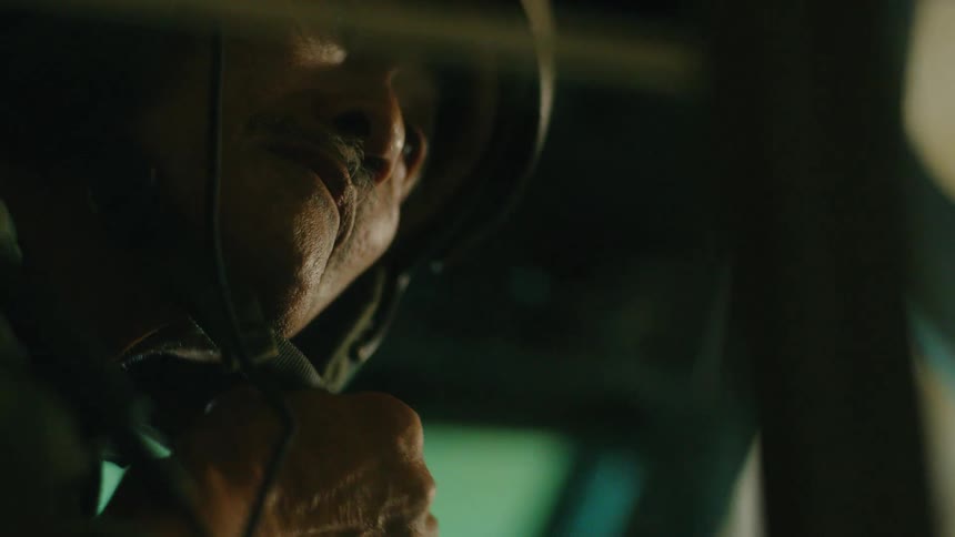 An older soldier with a mustache adjusts his helmet strap in the dim, atmospheric light of a military vehicle cockpit