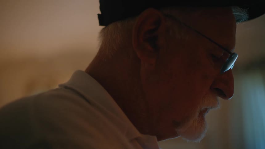 A silhouetted elderly man with glasses and a hat sits in a dark room, deeply focused on something just out of frame