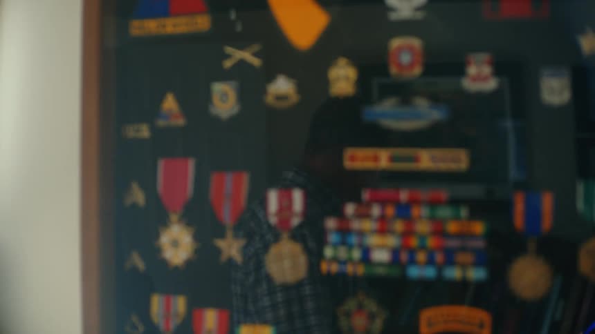 A male veteran's reflection is seen in the glass of his military shadow box filled with medals and ribbons before the focus shifts to the display