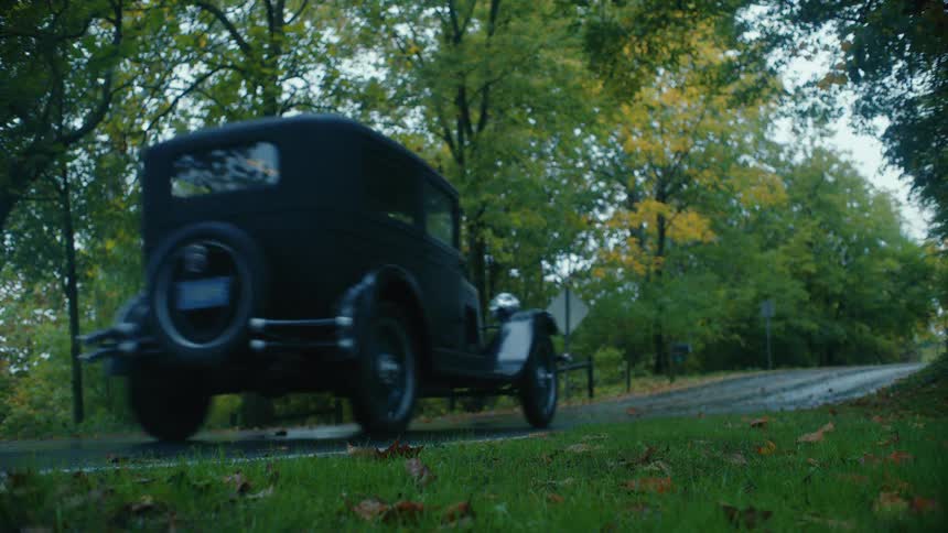 A vintage black 1920s-era car drives away down a wet, tree-lined road in a park during autumn