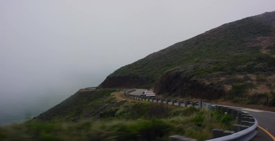 Driving along a winding coastal road on a foggy day, passing a classic car parked on the shoulder