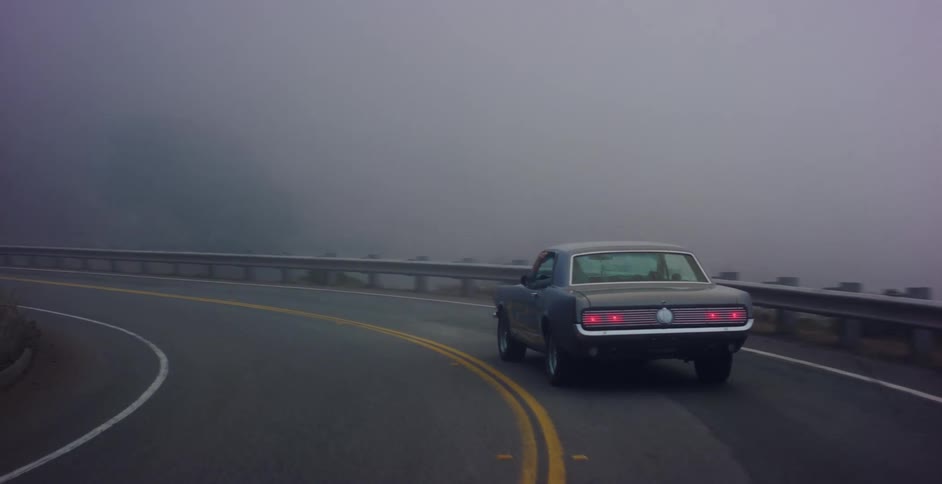 A vintage muscle car navigates a winding coastal highway enveloped in a thick, moody fog