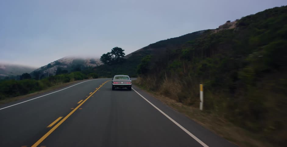 A vintage muscle car drives along a winding, two-lane road through foggy, tree-covered hills