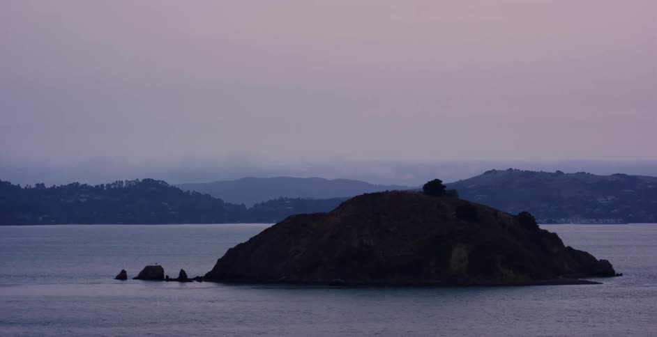 A silhouetted island rests in a calm bay with hazy mountains in the background during a moody twilight