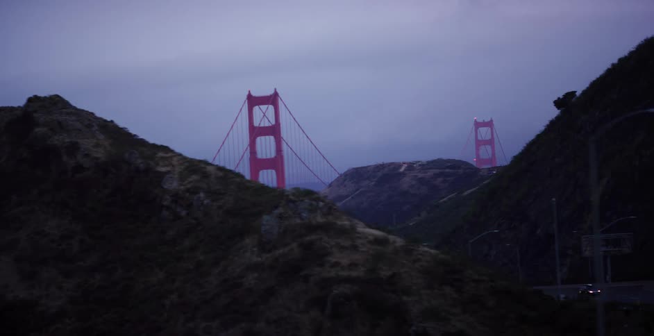 A moody driving shot reveals the iconic Golden Gate Bridge from behind rolling hills during a foggy dusk
