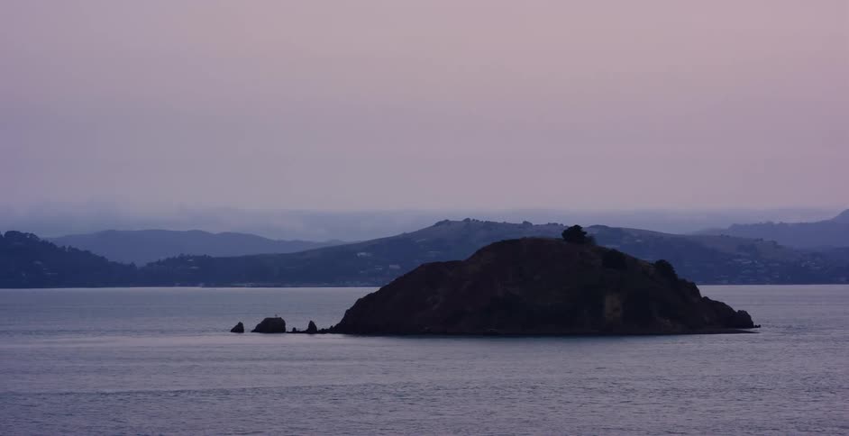 A solitary island silhouetted in the calm waters of a bay during a hazy, purple twilight