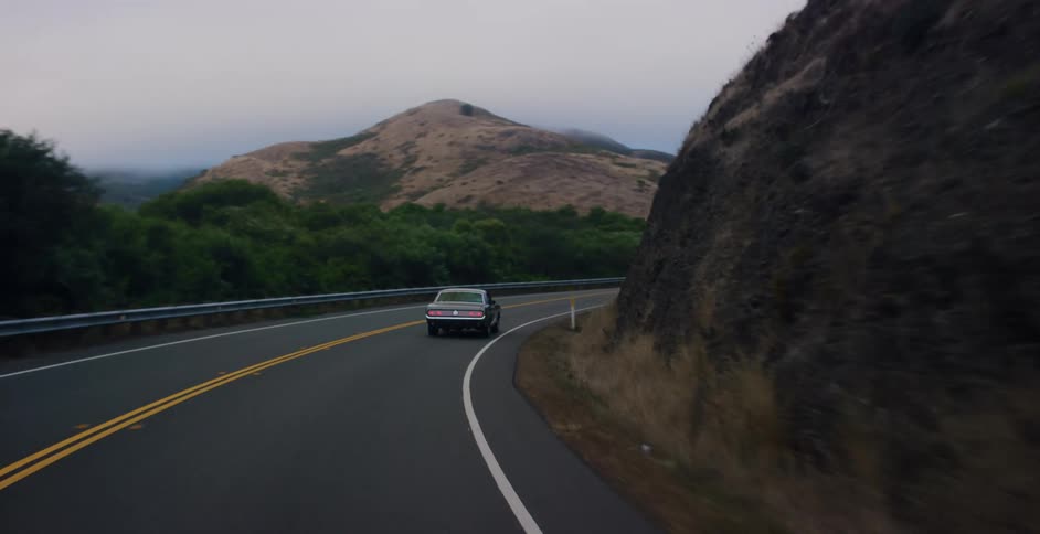 Following a classic American muscle car down a winding rural highway through foggy, rolling hills