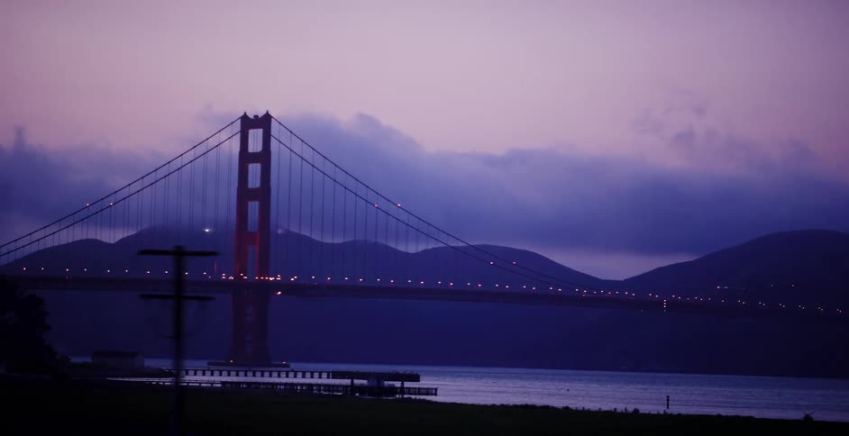 A moving shot reveals the iconic Golden Gate Bridge at dusk, with its lights glowing against a moody purple sky and silhouetted trees in the foreground