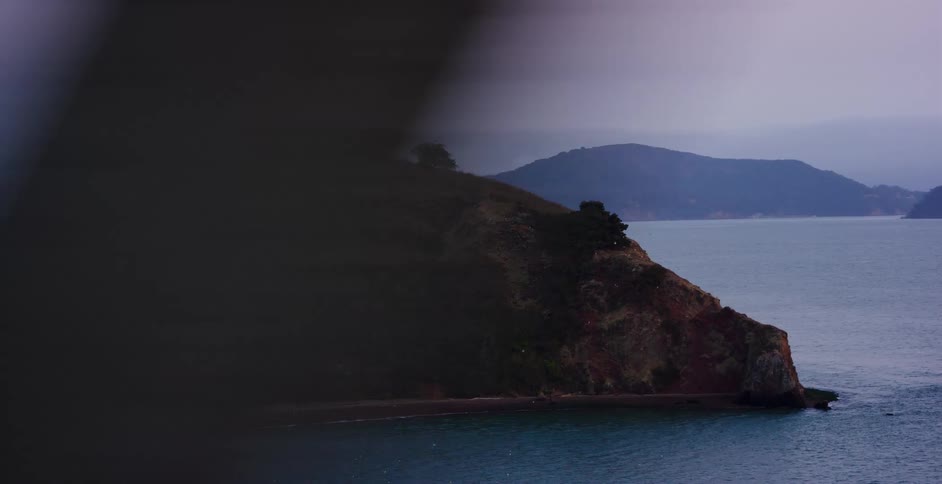 A small, tree-topped island rests in the calm ocean water on a grey, overcast evening