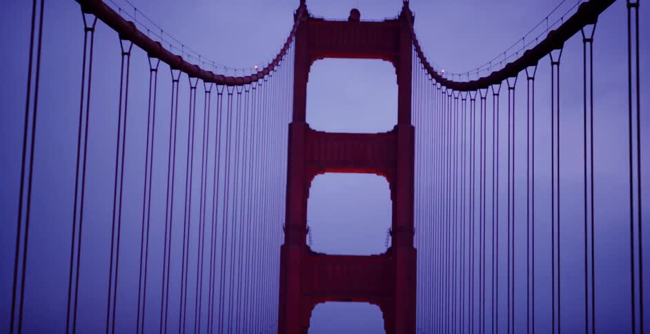 An upward tilting shot reveals the iconic red tower of the Golden Gate Bridge against a moody, blue twilight sky