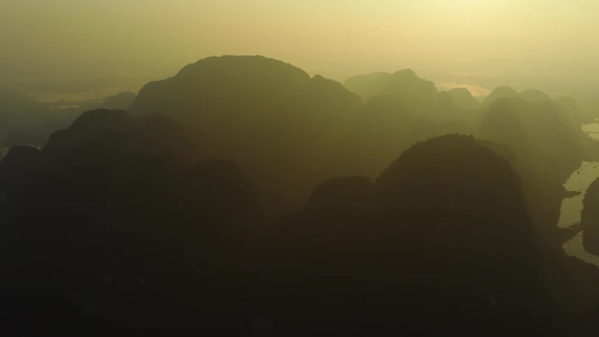 An atmospheric aerial shot reveals the silhouettes of a vast mountain range during a golden, hazy sunrise