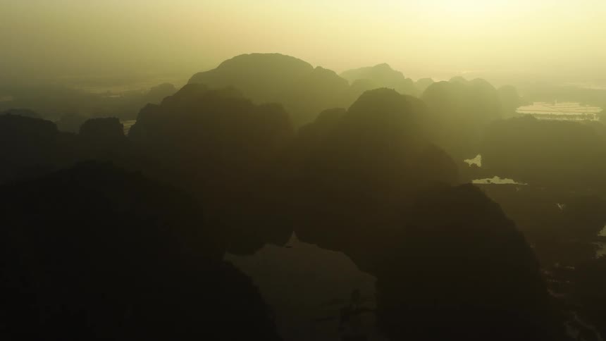 A slow aerial pullback reveals a vast, mystical landscape of silhouetted karst mountains shrouded in golden morning mist