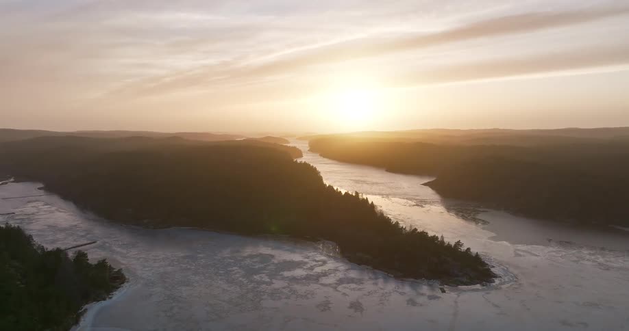 An expansive aerial shot slowly pulls back revealing a majestic winter sunrise over a frozen fjord surrounded by silhouetted, tree-covered hills