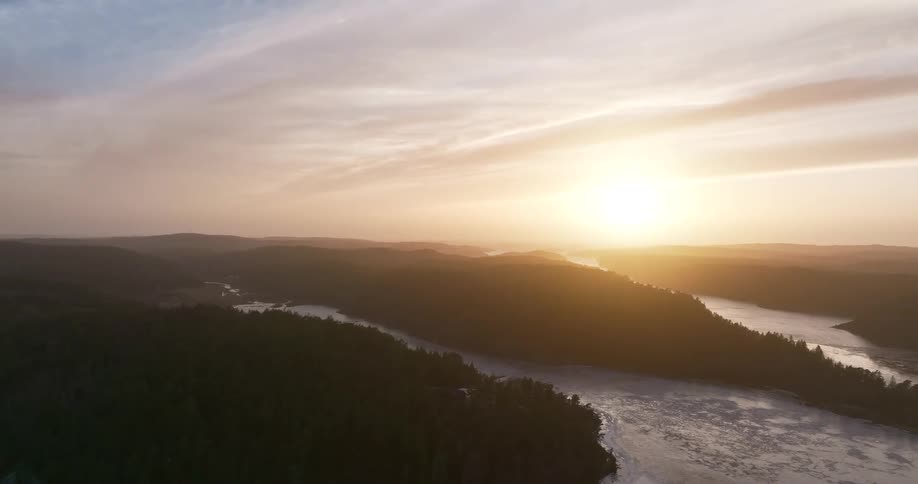 A stunning aerial view reveals a vast winter landscape with a golden sun rising over forested hills and a partially frozen river