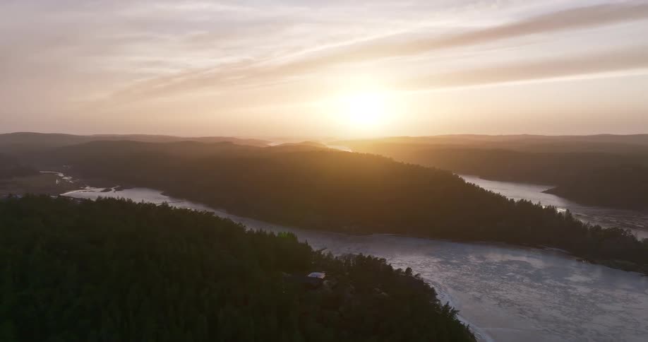 A serene aerial view shows the golden sun setting over a vast, hazy landscape of forested hills and a winding frozen river