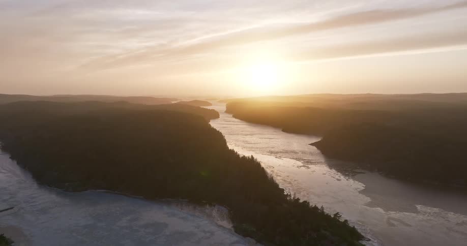 A serene aerial view shows the sun setting over a vast, wintry landscape with a partially frozen river winding through forested hills