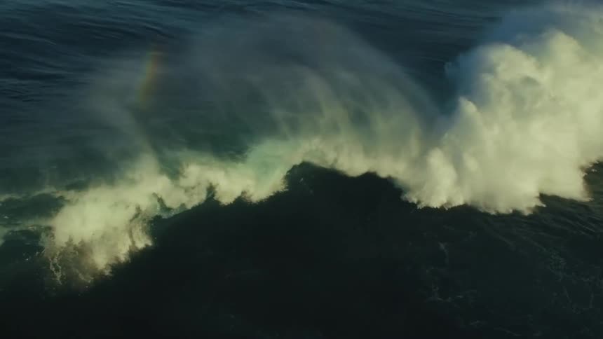 A massive ocean wave crests and breaks in dramatic slow motion, revealing a rainbow in its misty spray