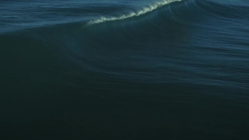 A gigantic, deep blue ocean wave rises and crashes in spectacular slow motion, its spray catching the light to form a rainbow