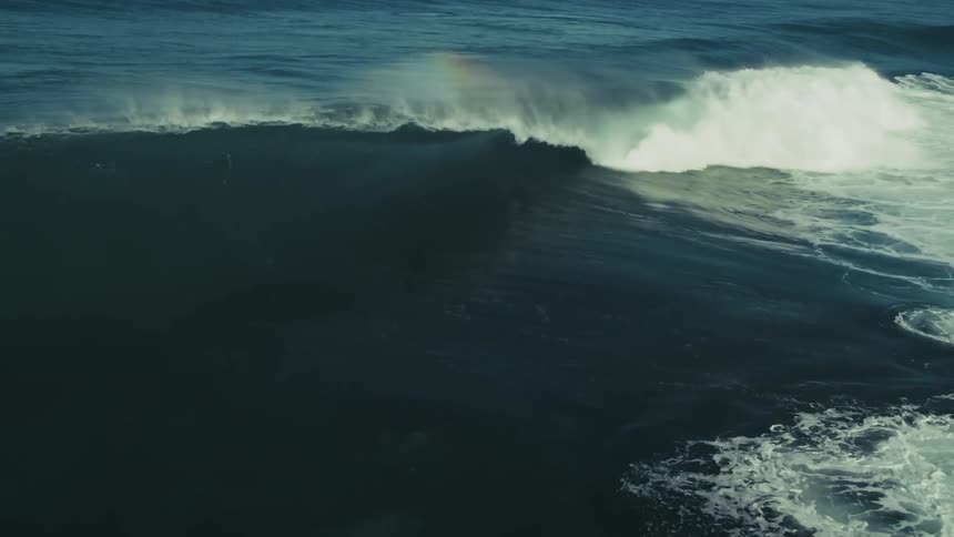 A massive deep blue ocean wave forms and crashes in dramatic slow motion, creating a rainbow in its misty spray