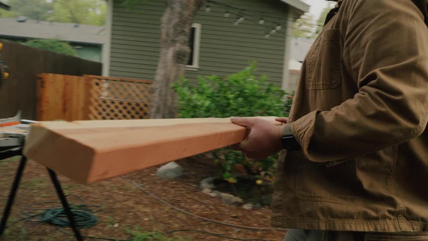 A handyman carries a long wooden beam across his backyard to a miter saw for a home improvement project in slow motion