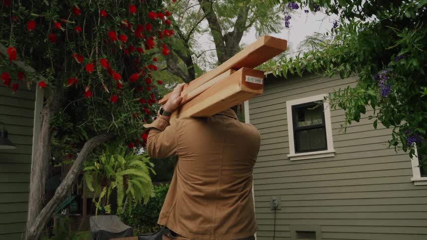 A man wearing a tool belt carries heavy wooden beams on his shoulder through his backyard in slow motion