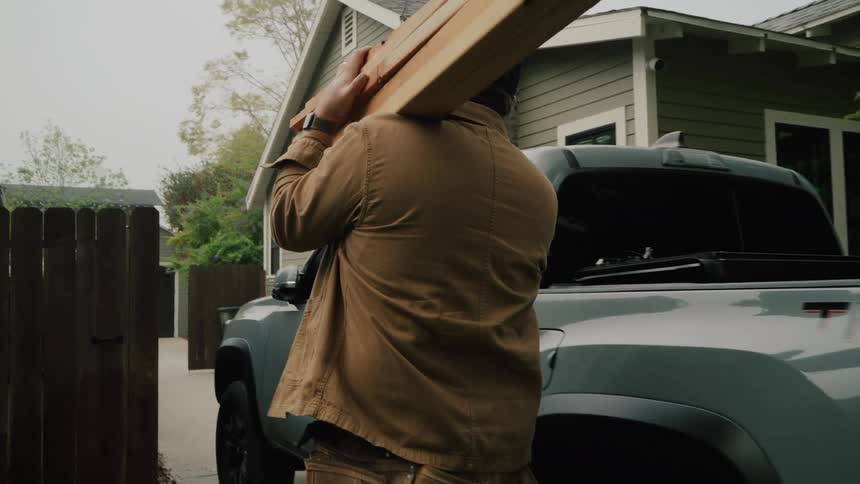 A handyman in slow motion carries a heavy stack of lumber from his truck into a residential yard