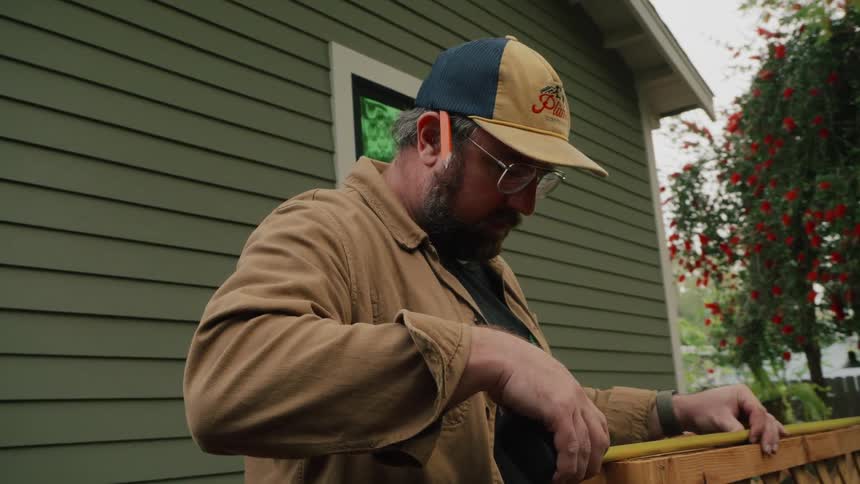 A man in a ball cap and glasses carefully measures a wooden lattice panel with a tape measure in his backyard