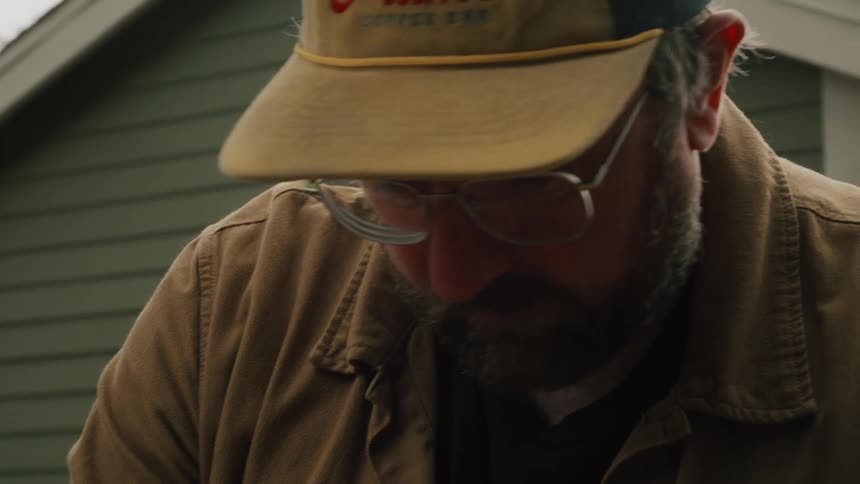 A bearded craftsman wearing a hat and glasses pulls a pencil from his cap to work on his project outdoors