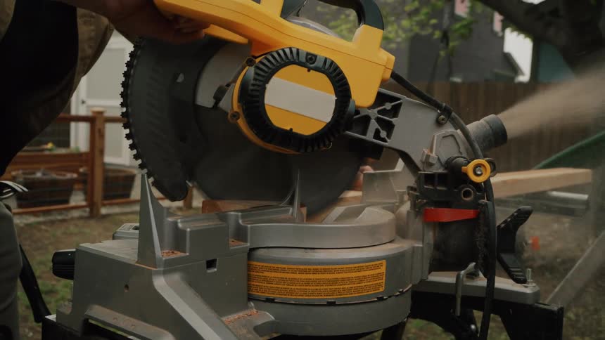 A carpenter wearing safety glasses carefully cuts a wood plank with a miter saw in his backyard, sending sawdust flying in slow motion