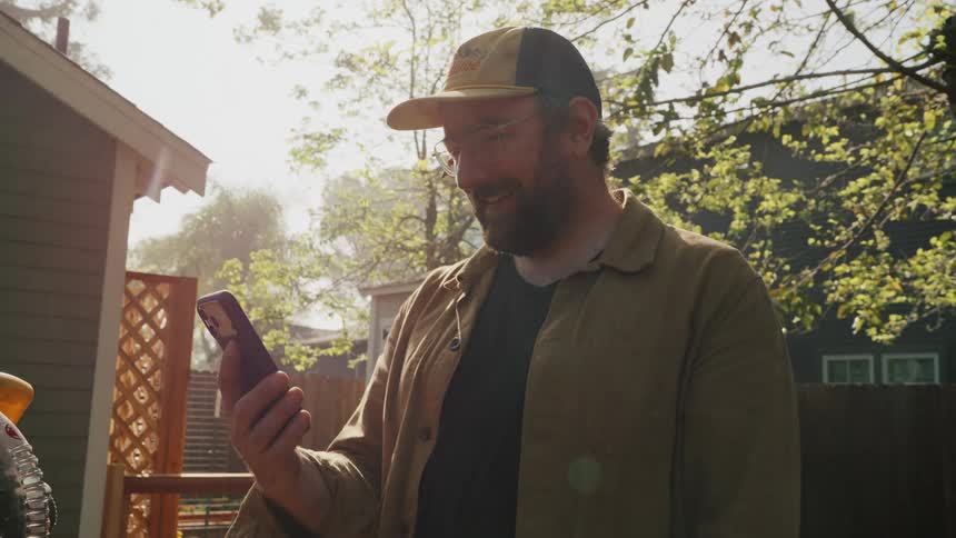 A smiling man with a beard and glasses looks at his smartphone in a sunny backyard