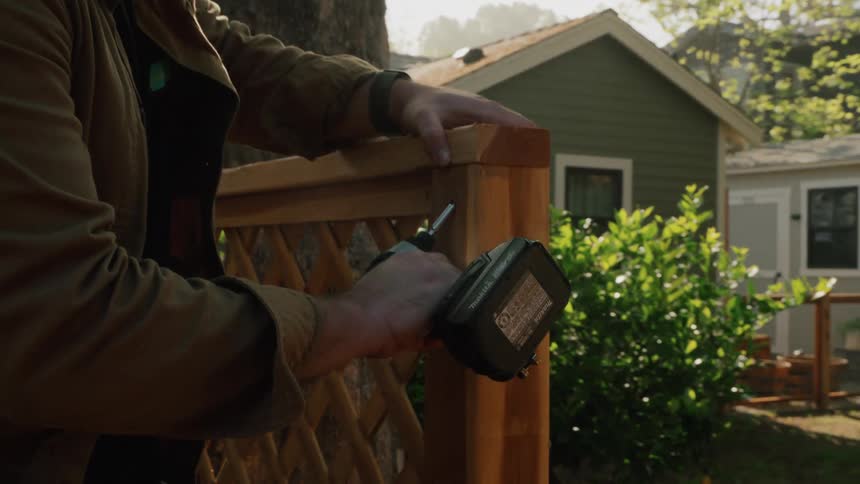 A focused handyman uses a power drill to assemble a new wooden lattice fence in a sunny residential backyard